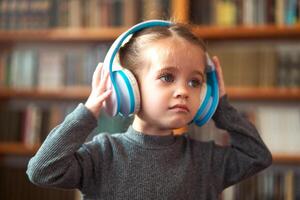 schattig Kaukasisch weinig meisje in groot wit hoofdtelefoons luistert muziek- in de interieur tegen de achtergrond van boekenplanken. foto