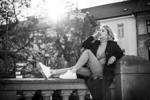 terug visie van vrouw in zwart jas en wit schoenen zittend Aan richel. elegant stedelijk mode portret. modieus vrouw model- genieten van stadsgezicht. gewoontjes buitenshuis levensstijl concept foto