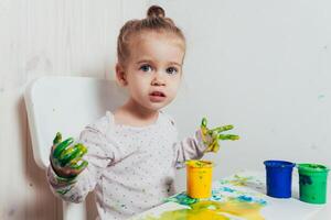 mooi weinig meisje trekt met vinger verven Aan een wit vel van papier. creatief kind ontwikkeling in kleuterschool of vrij tijd Bij huis foto