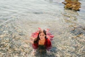 vrouw reizen zee. gelukkig toerist in rood jurk genieten nemen afbeelding buitenshuis voor herinneringen. vrouw reiziger poseren in zee strand, omringd door vulkanisch bergen, sharing reizen avontuur reis foto