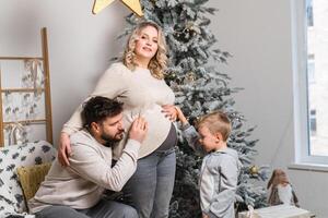 Kerstmis familie geluk portret van pa, zwanger mam en weinig zoon zittend fauteuil Bij huis in de buurt Kerstmis boom knuffel glimlach foto