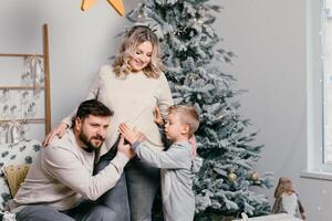 Kerstmis familie geluk portret van pa, zwanger mam en weinig zoon zittend fauteuil Bij huis in de buurt Kerstmis boom knuffel glimlach foto