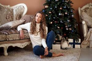 kerstmis. vrouw gekleed wit trui en jeans zittend Aan de verdieping in de buurt Kerstmis boom met Cadeau doos foto