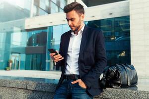 millennial zakenman met een mobiel telefoon in zijn handen. jong geslaagd bedrijf elegant Mens met een zwart leer zak wandelingen Aan een stad straat tegen de achtergrond van kantoor gebouwen foto