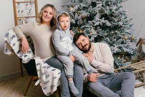 Kerstmis familie geluk portret van pa, zwanger mam en weinig zoon zittend fauteuil Bij huis in de buurt Kerstmis boom knuffel glimlach foto