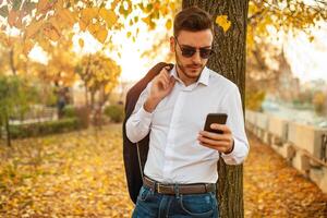 knap jong modieus Mens in een elegant bedrijf pak en zonnebril is typen een bericht in zijn telefoon wandelen in de herfst park Aan de achtergrond van helder geel bladeren. foto