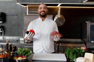 gelukkig glimlachen chef bereidt zich voor vlees schotel met divers groenten in de keuken. in een hand- de Mens houdt groenten, in de andere een stuk van vers vlees foto