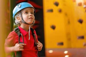 schattig weinig meisje klimmer in blauw beschermend helm en uitrusting voor beklimming staand in klimmer centrum amusement park voor kinderen foto