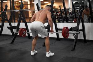 gespierd atletisch bodybuilder geschiktheid model- staand in voorkant van spiegel terug naar camera opleiding optillen barbell binnen- Sportschool foto