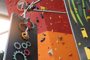 kind beklimming Aan muur in amusement centrum. beklimming opleiding voor kinderen. weinig meisje in gekleed beklimming uitrusting beklimmen hoog. extreem actief vrije tijd voor kinderen. foto