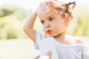 zonnig portret van een weinig kind drinken van een rietje sap Aan wazig zomer achtergrond foto