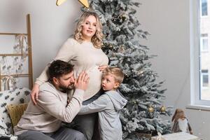 Kerstmis familie geluk portret van pa, zwanger mam en weinig zoon zittend fauteuil Bij huis in de buurt Kerstmis boom knuffel glimlach foto
