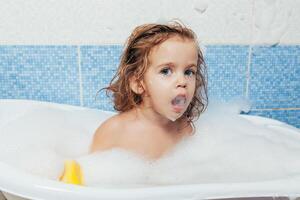 pret vrolijk gelukkig kleuter baby nemen een bad spelen met schuim bubbels. weinig kind in een bad. glimlachen kind in badkamer Aan blauw achtergrond. hygiëne en Gezondheid zorg. foto
