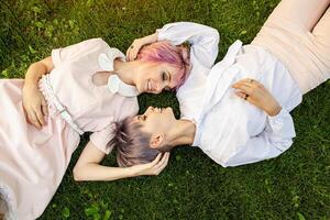 multiraciaal lesbienne paar aan het liegen Aan de gras. ze zijn twee jong Dames resting Bij park. een is Kaukasisch en de andere is aziatisch. ze zijn glimlachen en vervelend zomer kleren. foto