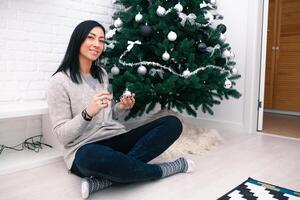 een jong vrouw zit in de buurt een Kerstmis boom, Holding een glas en een Kerstmis speelgoed. voorbereiding voor de nieuw jaar viering foto