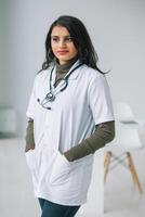 gelukkig jong Indisch vrouw dokter vervelend wit medisch jas en stethoscoop op zoek Bij camera. glimlachen vrouw arts poseren in ziekenhuis kantoor foto