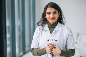 glimlachen vrouw arts poseren in ziekenhuis kantoor. gelukkig jong Indisch vrouw dokter vervelend wit medisch jas en stethoscoop op zoek Bij camera foto