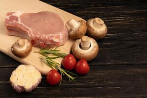 top visie van een stukken rauw varkensvlees karbonade steaks met kers tomaten rozemarijn champignon champignons en knoflook Aan een snijdend bord. foto