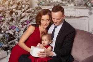 gelukkig jong familie met een kind Holding Kerstmis geschenk. zittend Aan de sofa in de buurt de Kerstmis boom Bij huis foto