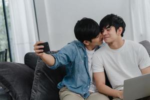 romantische jonge homo paar grappige selfie door mobiel thuis. aziatische minnaar man gelukkig ontspannen plezier met behulp van technologie mobiele telefoon glimlachend nemen een foto samen terwijl liggend bank in woonkamer concept.