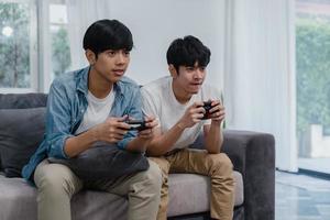 jong aziatisch homopaar speelt thuis spelletjes, tiener-koreaanse lgbtq-mannen met joystick hebben een grappig gelukkig moment samen op de bank in de woonkamer thuis. foto