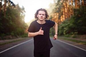 Kaukasisch jong gekruld Mens atleet loopt zonnig zomer dag Aan asfalt weg in de Woud. foto