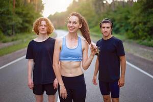 een groep van drie mensen atleten een meisje en twee mannen staand Aan asfalt weg in pijnboom Woud Bij zomer voordat jogging foto