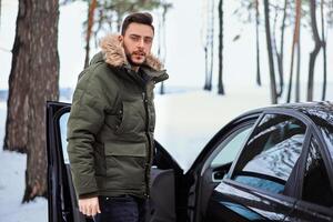 jong volwassen knap Mens staand in winter Woud in de buurt zijn auto. aantrekkelijk Kaukasisch vent aan het wachten voor helpen wanneer auto is gebroken. foto