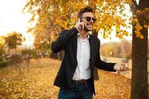 knap jong elegant mannetje zakenman gelukkig en glimlachen pratend Aan de telefoon Aan een wandelen in de herfst park Aan de achtergrond van geel bladeren foto