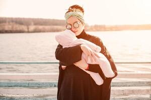 jong moeder draag- haar aanbiddelijk baby dochter in een baby vervoerder Aan mooi herfst park achter de meer foto