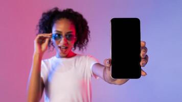 verrast zwart vrouw tonen smartphone met blanco scherm, neon backdrop foto