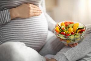 zwanger vrouw Holding kom met vers groente salat terwijl zittend Aan bed foto