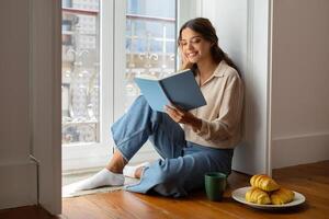 glimlachen jong vrouw lezing boek terwijl comfortabel zittend Aan verdieping in de buurt venster foto