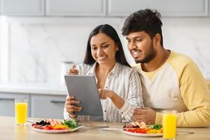 vrolijk Indisch echtgenoten gebruik makend van digitaal tablet terwijl aan het eten ontbijt foto