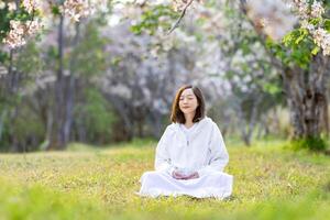 Aziatisch vrouw is aan het doen meditatie onder bloem boom gedurende kers bloeiende seizoen voor binnenste vrede, opmerkzaamheid en zen praktijk foto