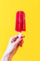 ai gegenereerd hand- Holding een levendig rood ijslolly tegen een helder geel achtergrond foto