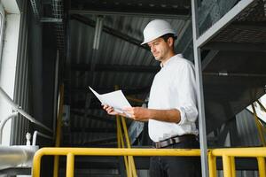 een jong geslaagd ingenieur met een tekening in zijn handen is staand in de gebied van een modern fabriek. foto