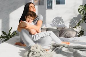 jong moeder met haar 5 jaren oud weinig zoon gekleed in pyjama zijn ontspannende en spelen in de bed Bij de weekend samen, lui ochtend, warm en knus tafereel. familie huis schieten. foto