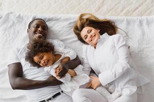 portret van gelukkig multiraciaal jong familie aan het liegen Aan knus wit bed Bij huis, glimlachen Internationale mam en vader ontspannende met weinig biraciaal meisje kind poseren voor afbeelding in slaapkamer foto