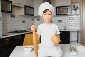 jongen in koken hoed zittend Bij een keuken balie. weinig jongen in keuken foto