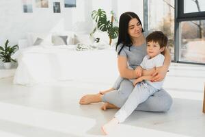 jong moeder met haar 4 jaren oud weinig zoon gekleed in pyjama zijn ontspannende en spelen in de bed Bij de weekend samen, lui ochtend, warm en knus tafereel. foto