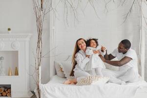 portret van gelukkig multiraciaal jong familie aan het liegen Aan knus wit bed Bij huis, glimlachen Internationale mam en vader ontspannende met weinig biraciaal meisje kind poseren voor afbeelding in slaapkamer foto