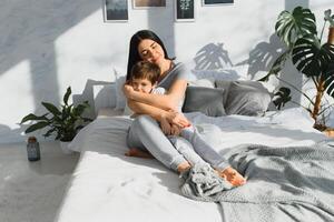 jong moeder met haar 5 jaren oud weinig zoon gekleed in pyjama zijn ontspannende en spelen in de bed Bij de weekend samen, lui ochtend, warm en knus tafereel. familie huis schieten. foto