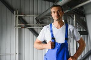 jong glimlachen professioneel in overall en beschermend helm staand in voorkant van camera binnen groot machine bouwen fabriek foto