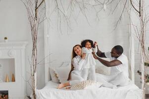 portret van gelukkig multiraciaal jong familie aan het liegen Aan knus wit bed Bij huis, glimlachen Internationale mam en vader ontspannende met weinig biraciaal meisje kind poseren voor afbeelding in slaapkamer foto