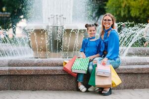 mooi mam en haar schattig weinig dochter zijn Holding boodschappen doen Tassen, op zoek Bij camera en glimlachen terwijl staand buitenshuis. boodschappen doen concept. foto