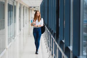 vrolijk brunette leerling meisje met zwart rugzak houdt boeken in modern gebouw. vrouw leerling staand met boeken in college gang foto