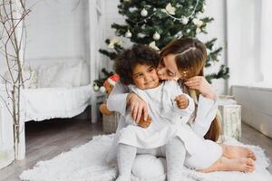 vrolijk Kerstmis en gelukkig vakantie vrolijk mam en haar schattig dochter Bij Kerstmis boom. ouder en weinig kind hebben pret in de buurt Kerstmis boom binnenshuis. liefhebbend familie met presenteert in kamer. foto