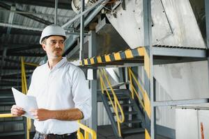 een jong geslaagd ingenieur met een tekening in zijn handen is staand in de gebied van een modern fabriek. foto