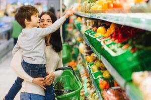 vrouw en kind jongen gedurende familie boodschappen doen met trolley Bij supermarkt foto